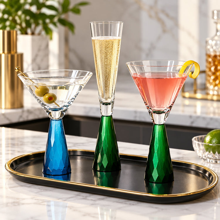 Three elegant stemmed cocktail glasses with faceted colored bases are arranged on a black tray with gold trim. One glass is filled with a clear drink, one with a pale gold bubbly drink, and one with a pink drink and a lemon twist.
