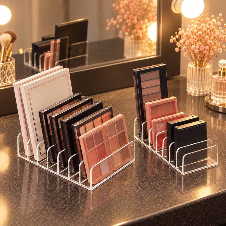 Clear acrylic makeup organizer stands on a dark countertop, holding multiple eyeshadow palettes and compact powders. A mirror and warm vanity lights reflect the setup, with decorative flowers and a perfume bottle in the background.