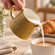 A hand holds a textured gold milk pitcher with a white interior, pouring milk into a cup of coffee. A croissant is blurred in the background on a table with a folded cloth.