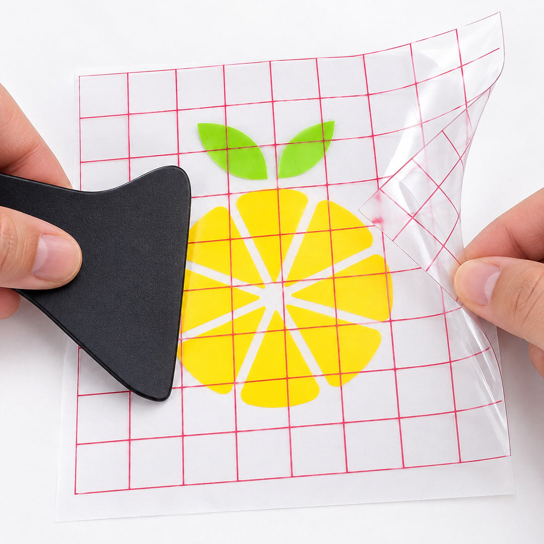 A person's hands peel a clear vinyl transfer sheet from a backing, using a black squeegee to smooth the sheet. The transfer features a yellow citrus slice design with green leaves, set against a white background with a red grid pattern.