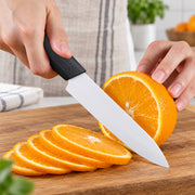 A kitchen knife with a black textured handle and a silver blade is slicing a fresh orange on a wooden cutting board, with a person's hands holding the fruit and knife in a bright kitchen setting.