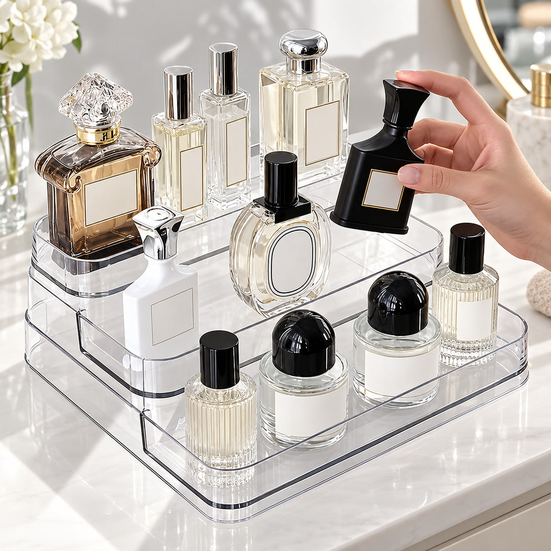 A hand holds a black perfume bottle next to a clear acrylic organizer filled with various perfume and cosmetic bottles on a white vanity counter.