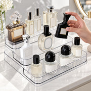 A hand holds a black perfume bottle next to a clear acrylic organizer filled with various perfume and cosmetic bottles on a white vanity counter.