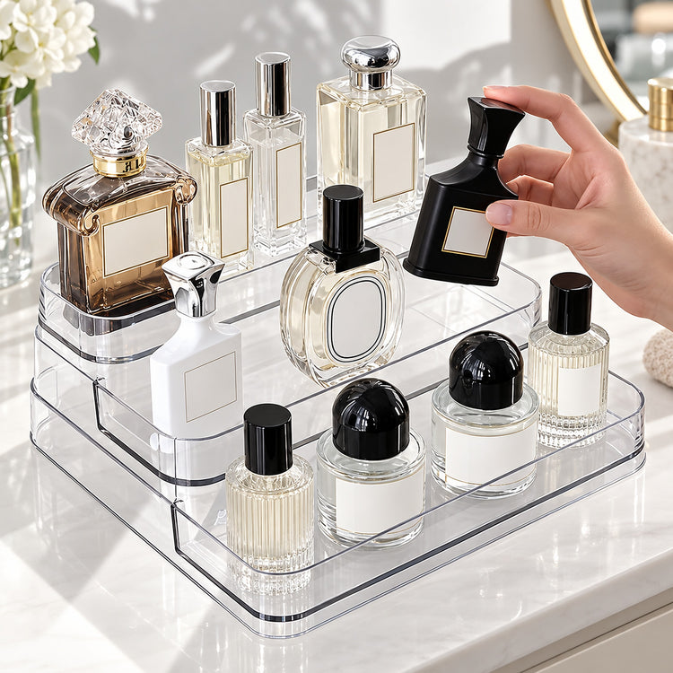 A hand holds a black perfume bottle next to a clear acrylic organizer filled with various perfume and cosmetic bottles on a white vanity counter.