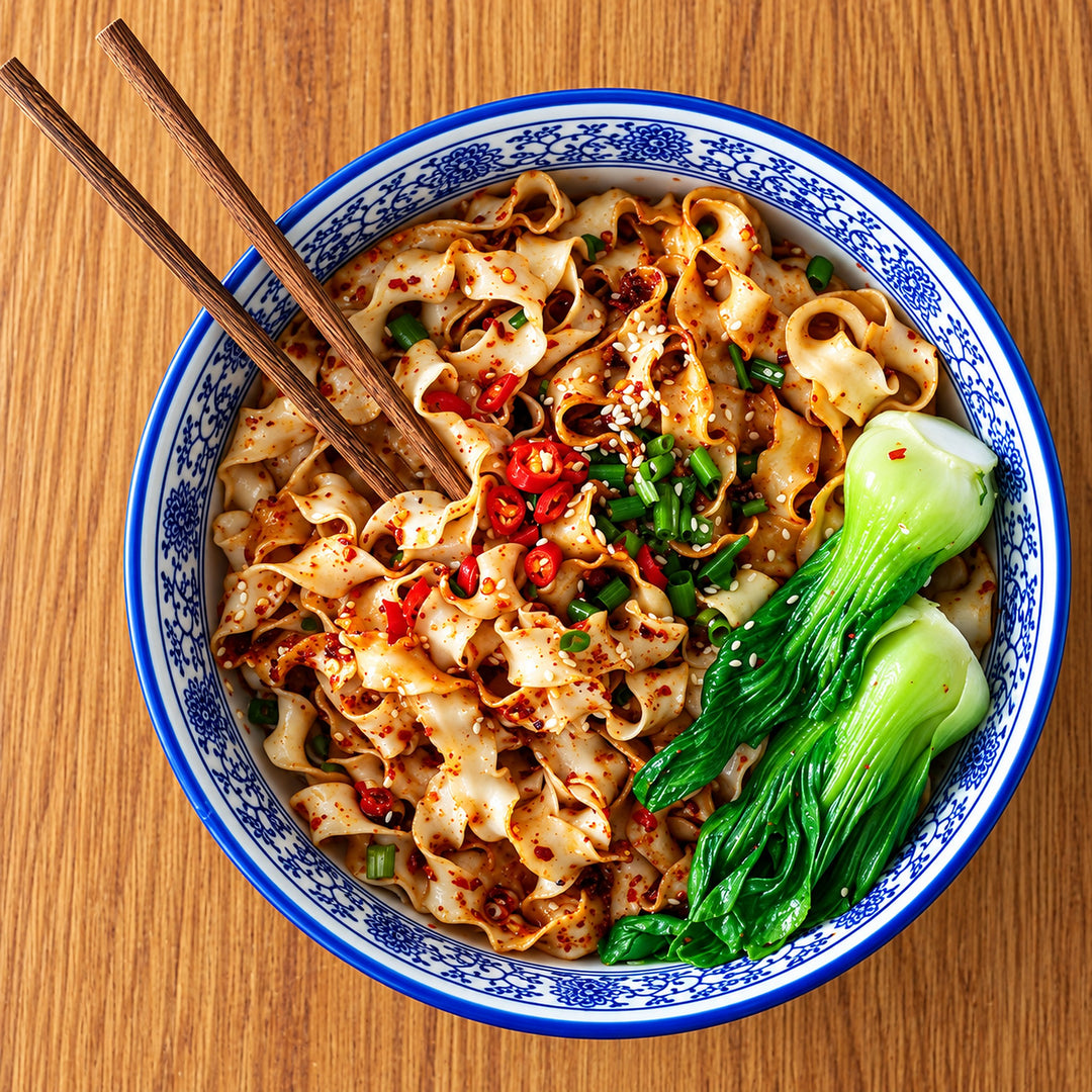 A top-down view of a bowl of spicy flat noodles with red chili oil, chopped green onions, and two pieces of bok choy on a wooden table, with wooden chopsticks resting on the side.