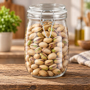A clear glass clamp-lid jar filled with shelled pistachios, set on a rustic wooden table with a softly blurred kitchen background.