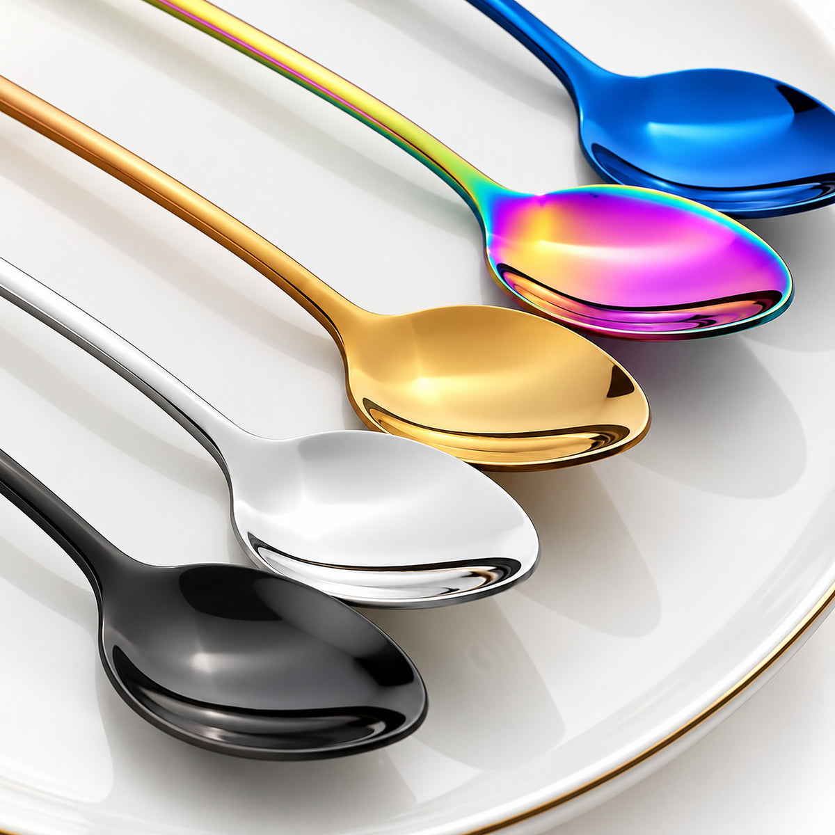 Four stainless steel spoons with different finishes arranged diagonally on a white plate: black, silver, gold, and a rainbow iridescent blue.