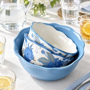 A set of nested ceramic bowls with a scalloped rim, featuring a blue floral and leaf pattern on a white base, nested inside a larger solid light blue bowl. The bowls are styled on a white tablecloth with glassware, lemon slices, and a vase of flowers in the background.