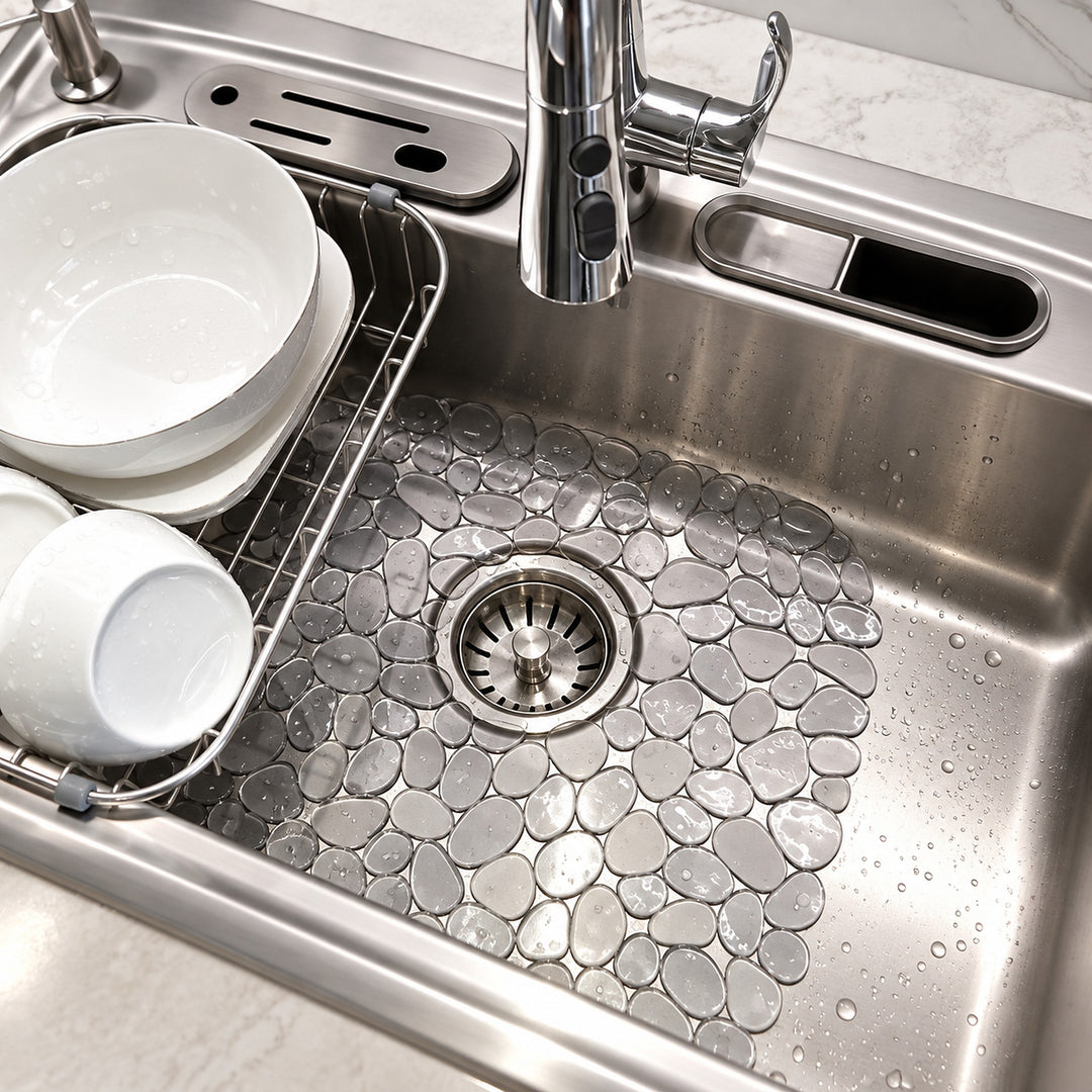A close-up of a stainless steel kitchen sink with a textured, pebble-patterned drain cover. White ceramic dishes are stacked on the left inside a wire drying rack, and a chrome faucet is visible at the top.