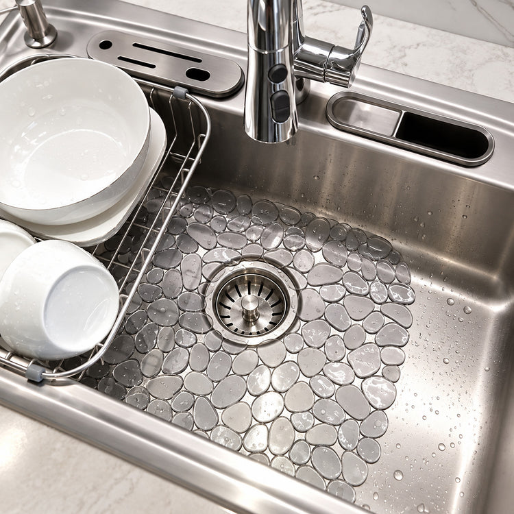 A close-up of a stainless steel kitchen sink with a textured, pebble-patterned drain cover. White ceramic dishes are stacked on the left inside a wire drying rack, and a chrome faucet is visible at the top.