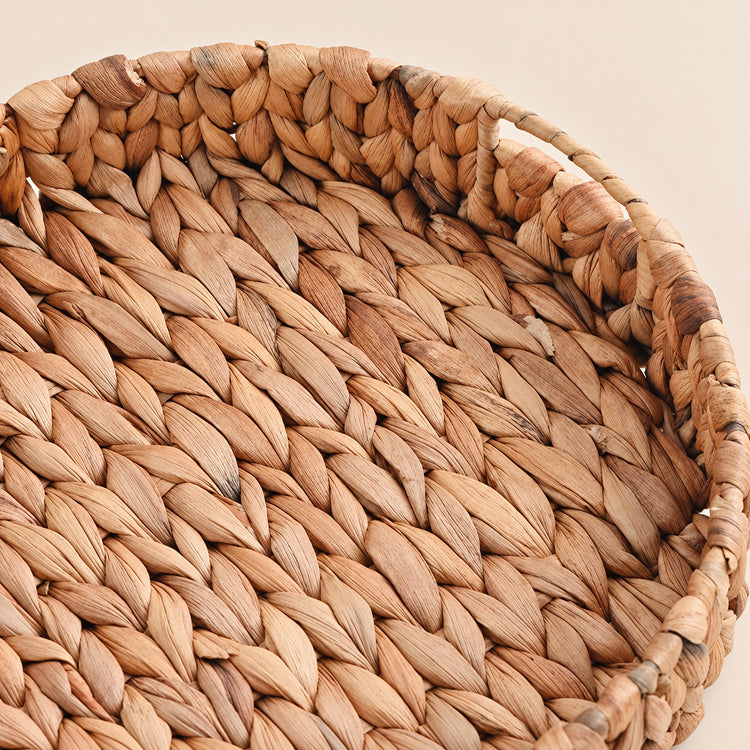 Close-up of a round woven basket made from natural tan and brown seaweed or straw fibers, showing the detailed braided texture and handle on a light beige background.