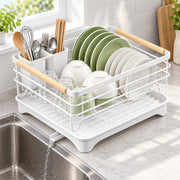 A white plastic and metal dish rack with light wood handles, shown holding green and white plates, bowls, and utensils in a bright kitchen setting above a sink.