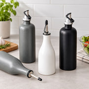 Four matte-finish oil or vinegar bottles with pour spouts arranged on a light countertop in a kitchen setting. The set includes two gray bottles, one white bottle, and one black bottle, with one gray bottle lying on its side in the foreground.