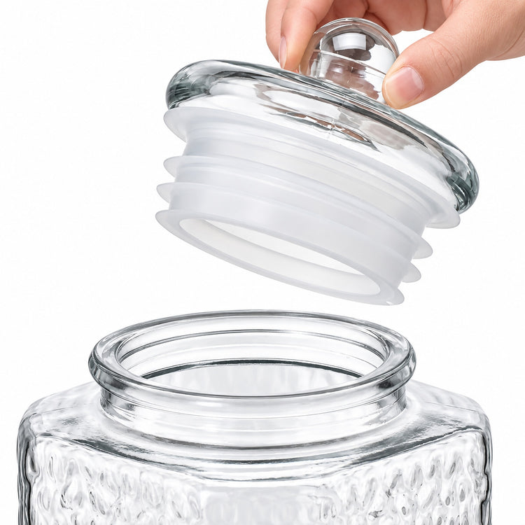 A clear glass jar with a textured exterior and a matching glass lid being lifted by a hand, showing a white silicone gasket ring between the glass pieces on a white background.
