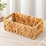 A rectangular woven straw basket with integrated handles, shown on a light wood tabletop with soft neutral curtains and a small plant in the background.
