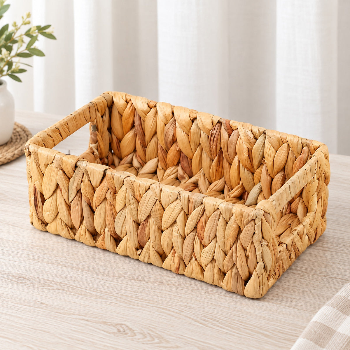A rectangular woven straw basket with integrated handles, shown on a light wood tabletop with soft neutral curtains and a small plant in the background.