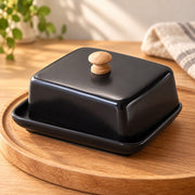 A glossy black ceramic butter dish with a matching square lid and a light wood knob, set on a round wooden tray in a softly lit kitchen setting.