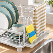 A white plastic and metal dish drying rack on a kitchen counter, holding stacked plates, a cup, wine glasses, a yellow sponge with a blue band, and a beige-and-white striped towel. A sink is visible in the foreground.