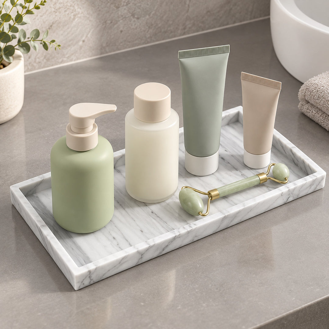 A set of minimalist bathroom or skincare containers arranged on a white marble tray, including a sage green pump bottle, a frosted white bottle, a sage green tube, a beige squeeze tube, and a jade roller, on a gray countertop.