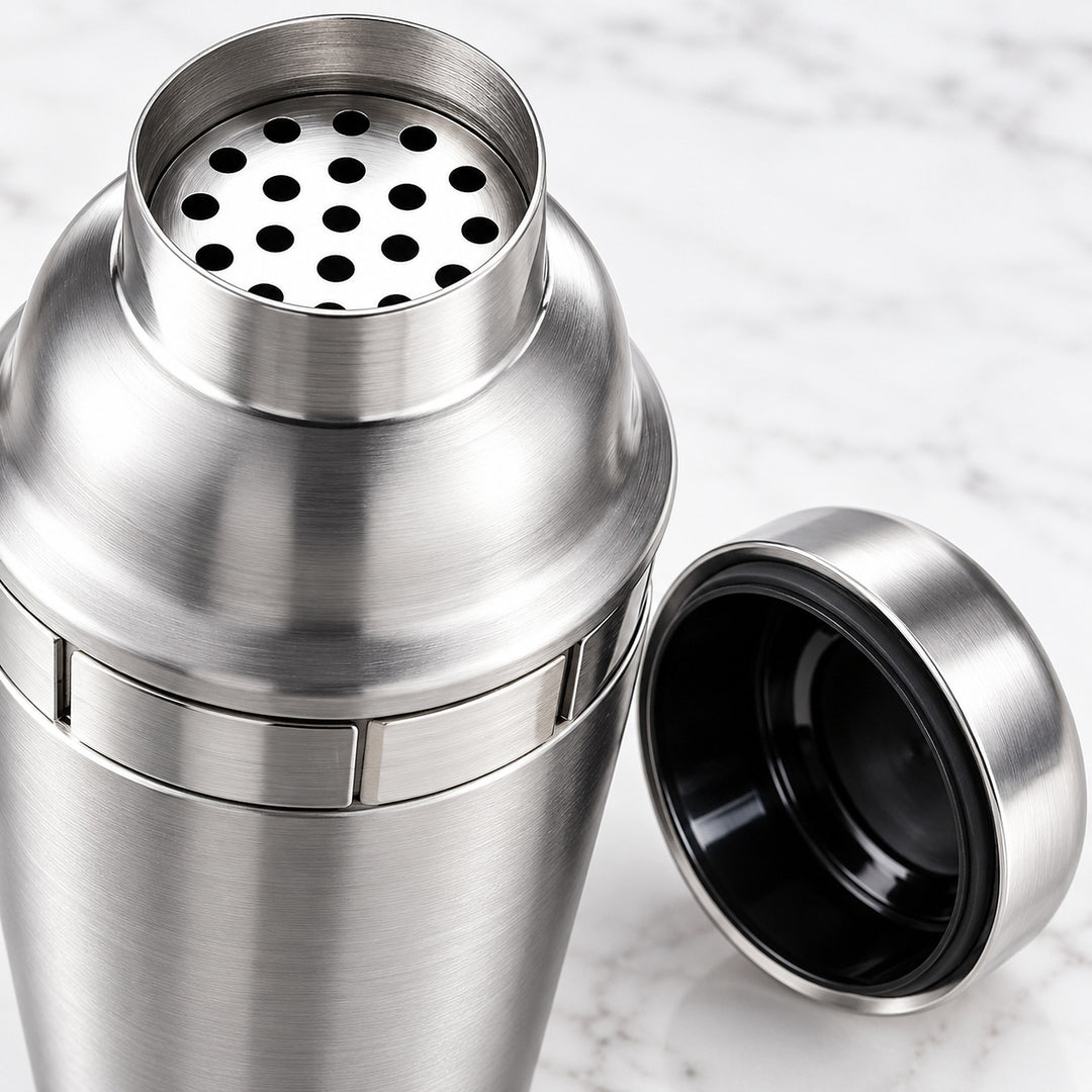 A stainless steel cocktail shaker with its lid off, showing the perforated top and black interior, set against a light marble background.