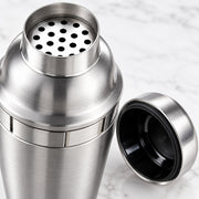 A stainless steel cocktail shaker with its lid off, showing the perforated top and black interior, set against a light marble background.