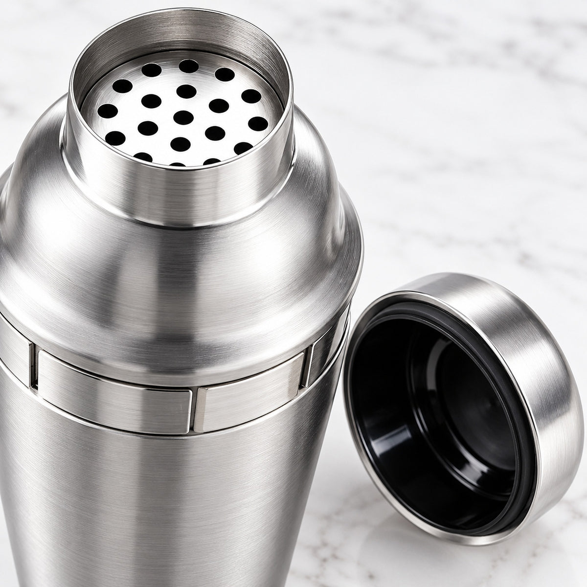 A stainless steel cocktail shaker with its lid off, showing the perforated top and black interior, set against a light marble background.