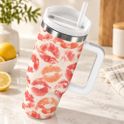 A handled travel tumbler with a white lid and straw, covered in a pink and orange lip print pattern, set on a marble countertop with lemons and a plant in the background.