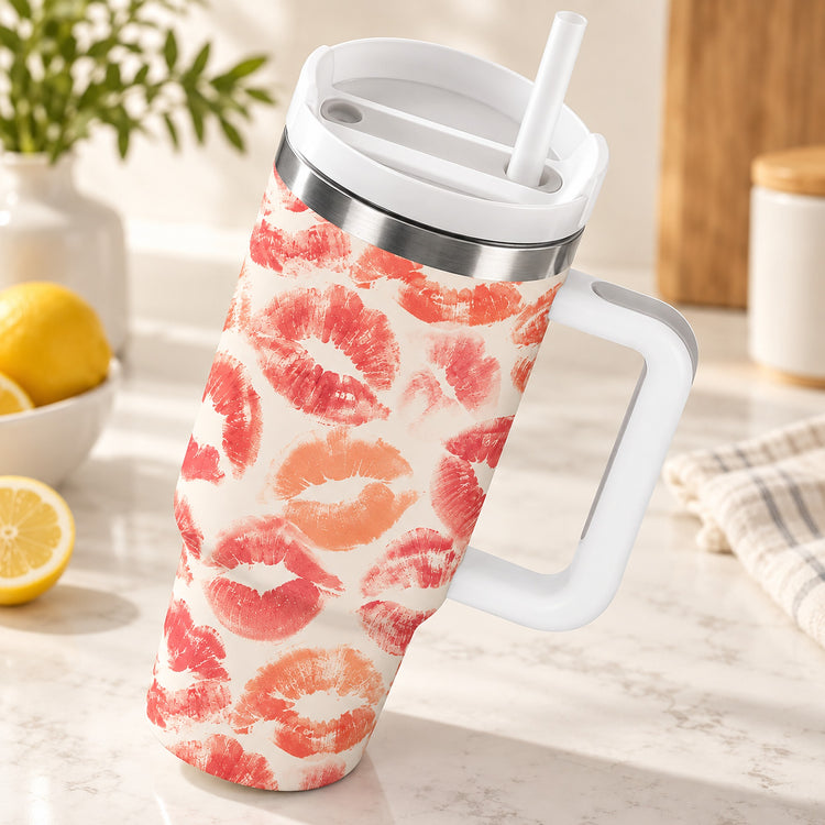 A handled travel tumbler with a white lid and straw, covered in a pink and orange lip print pattern, set on a marble countertop with lemons and a plant in the background.