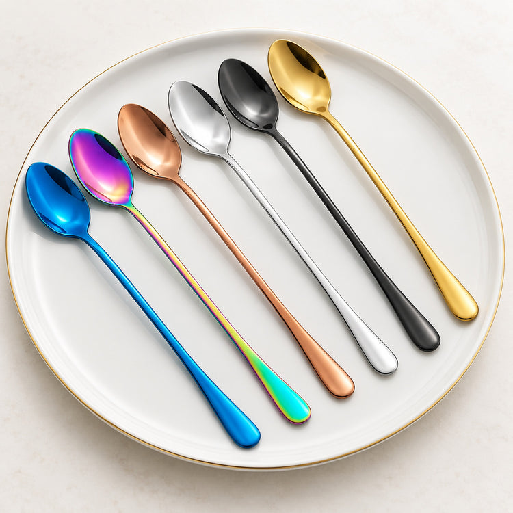 Six stainless steel spoons in different metallic colors are arranged diagonally on a white ceramic plate with a gold rim. The spoons are blue, iridescent rainbow, rose gold, silver, black, and gold.