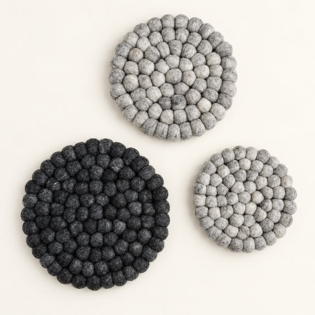 Three round felt coasters made of small textured pom-pom circles, shown on a plain light background. Two coasters are dark charcoal/black and one is light gray.