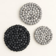 Three round felt coasters made of small textured pom-pom circles, shown on a plain light background. Two coasters are dark charcoal/black and one is light gray.