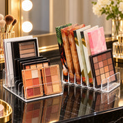 Clear acrylic makeup organizer stands on a glossy black marble vanity surface, holding eyeshadow palettes and colorful makeup booklets in a bright vanity setting with mirror lights and flowers in the background.