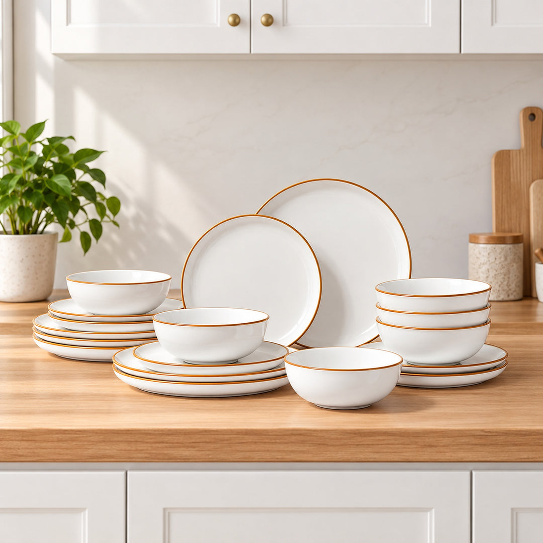 A coordinated dinnerware set arranged on a wooden kitchen counter, including white plates and bowls with thin gold rims. White cabinets, a potted plant, and kitchen accessories are visible in the background.