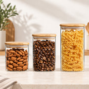 Three clear glass canisters with light wood lids, filled with almonds, coffee beans, and rotini pasta, arranged on a light countertop in a bright kitchen setting.