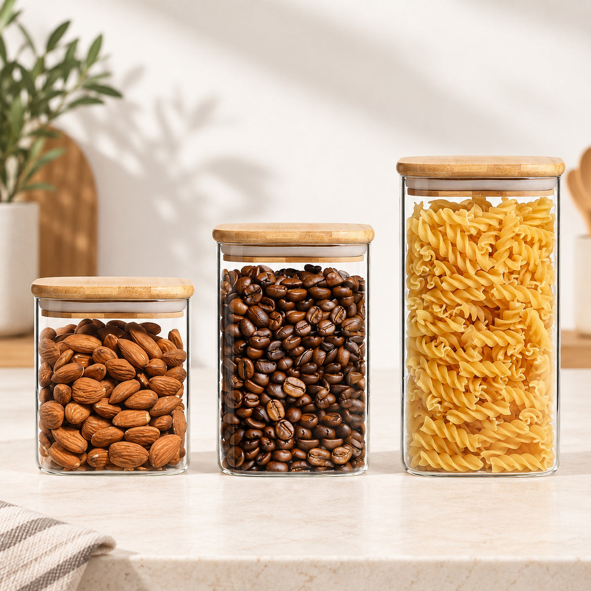 Three clear glass canisters with light wood lids, filled with almonds, coffee beans, and rotini pasta, arranged on a light countertop in a bright kitchen setting.