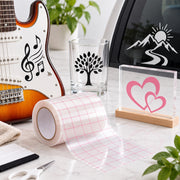 A roll of clear transfer tape with a pink grid pattern is unrolled on a light marble surface. Surrounding it are example applications: a clear glass tumbler with a black tree design, a clear acrylic block with pink heart designs on a wooden base, and a guitar with black musical note and treble clef decals.