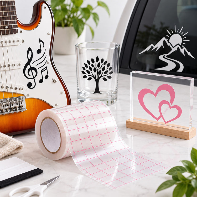 A roll of clear transfer tape with a pink grid pattern is unrolled on a light marble surface. Surrounding it are example applications: a clear glass tumbler with a black tree design, a clear acrylic block with pink heart designs on a wooden base, and a guitar with black musical note and treble clef decals.