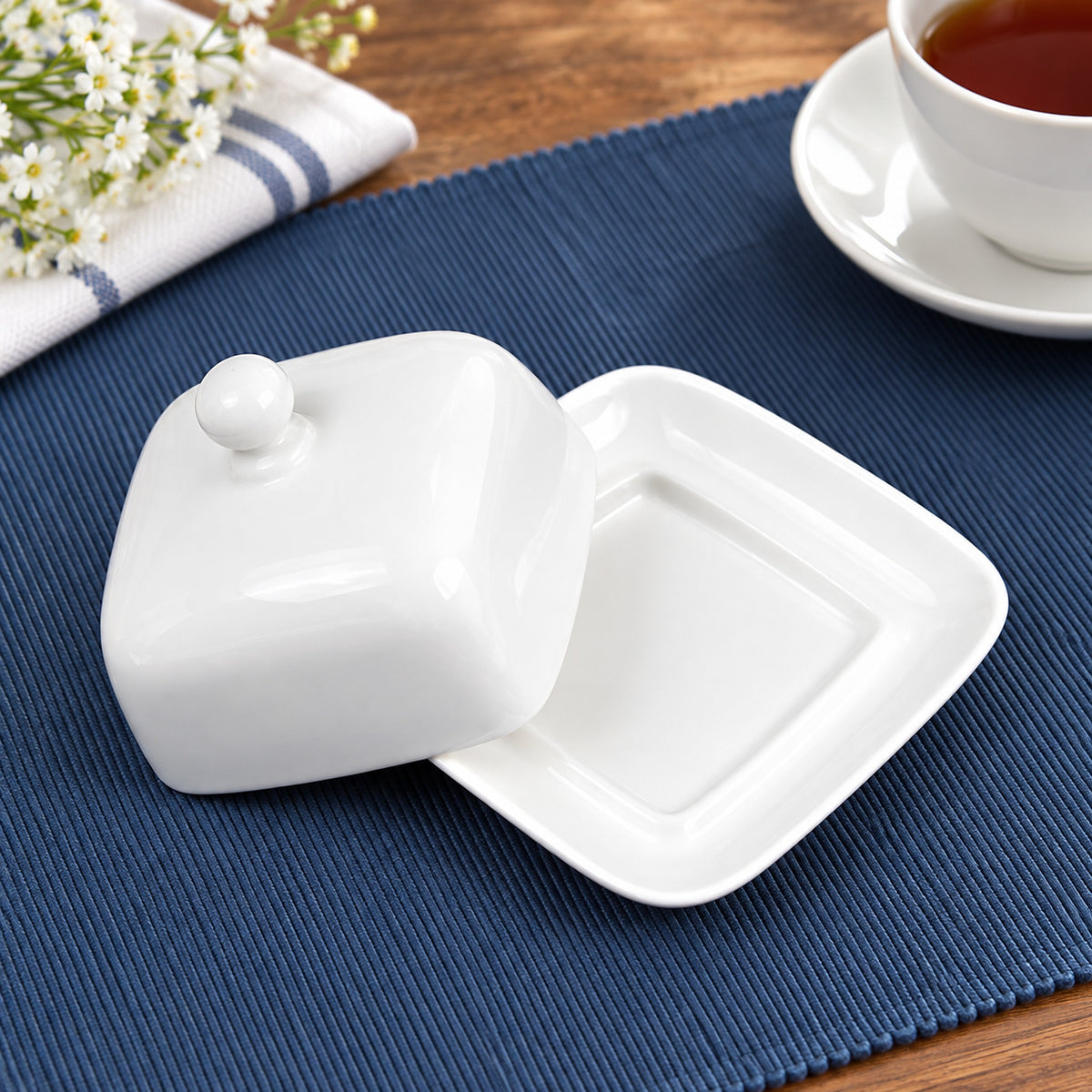 A white ceramic butter dish with a matching lid, shown on a blue textured placemat with a cup of tea and flowers in the background.