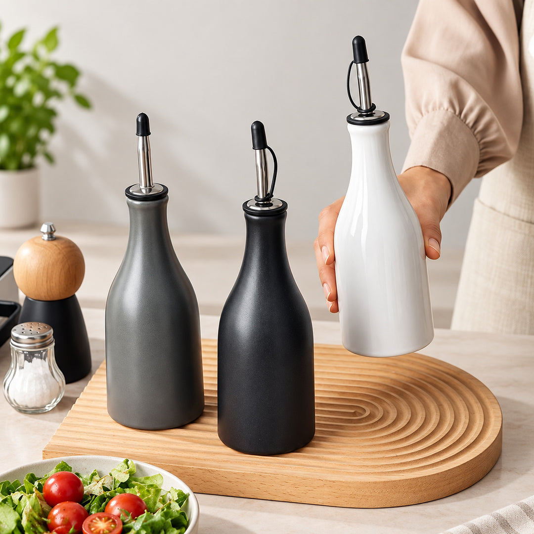 Three matte bottle-style oil or vinegar dispensers with pour spouts are arranged on a round wooden board in a kitchen setting. One dispenser is being held by a person whose arm and torso are partially visible on the right.