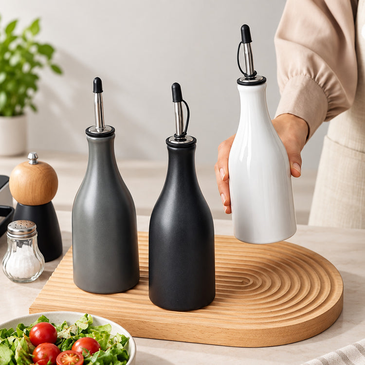Three matte bottle-style oil or vinegar dispensers with pour spouts are arranged on a round wooden board in a kitchen setting. One dispenser is being held by a person whose arm and torso are partially visible on the right.
