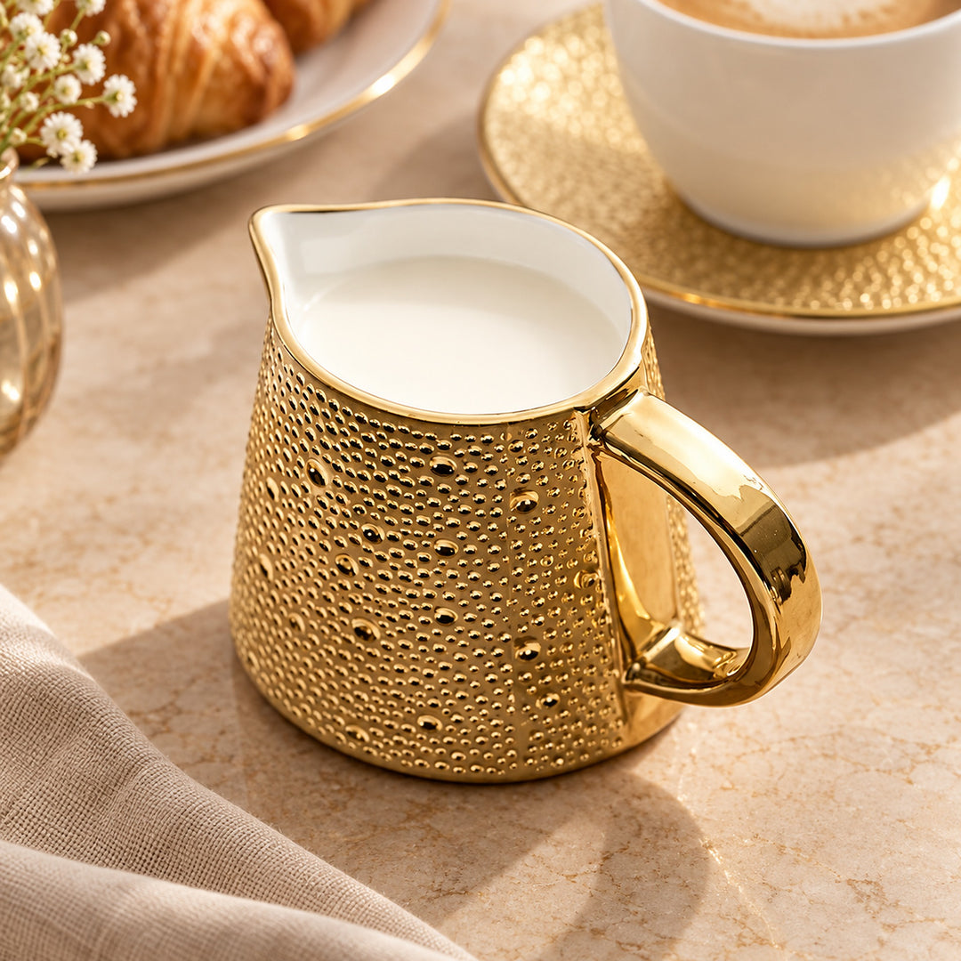 A gold ceramic milk pitcher with a textured, hammered surface and a white interior, filled with milk, set on a beige stone surface with a cup and croissant in the background.