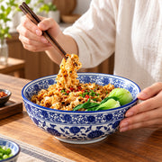 A person holding chopsticks over a blue-and-white patterned bowl of noodles with vegetables, seated at a wooden table.
