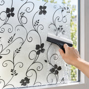 A hand holds a squeegee against a frosted glass window with a black floral scroll pattern applied, showing the textured, wet surface. A white and black window squeegee is shown applying a decorative frosted film or privacy glass to a window.
