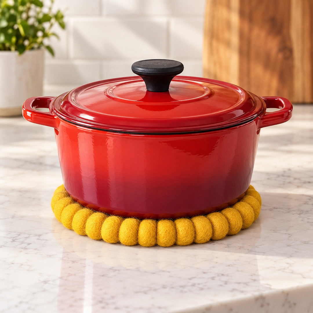 A red enameled cast iron pot with a matching lid and black knob, sitting on a yellow felt trivet on a white marble kitchen countertop.