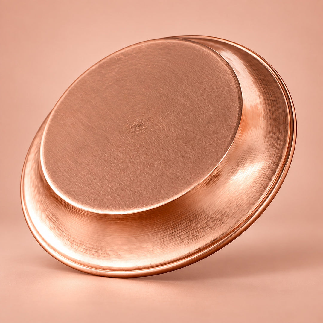 A hammered copper-colored metal tray or shallow pan shown at an angle on a warm peach-pink studio background.
