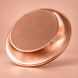 A hammered copper-colored metal tray or shallow pan shown at an angle on a warm peach-pink studio background.
