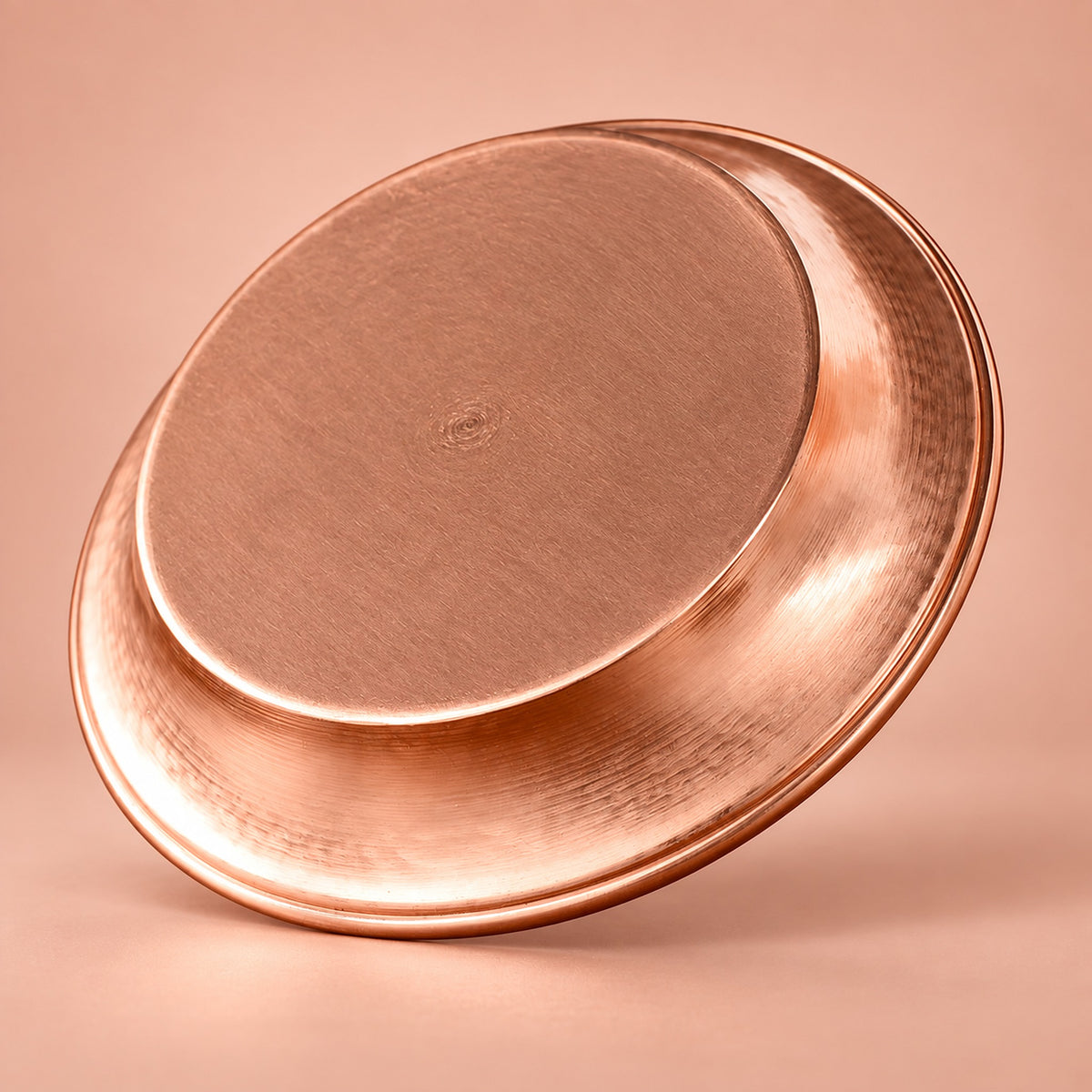 A hammered copper-colored metal tray or shallow pan shown at an angle on a warm peach-pink studio background.