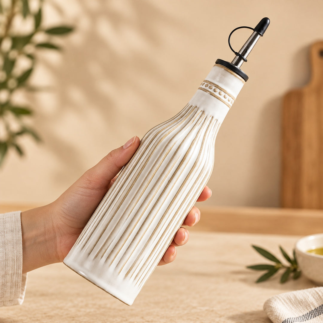 A hand holds a ribbed ceramic oil bottle with a black pour spout against a warm beige kitchen background.