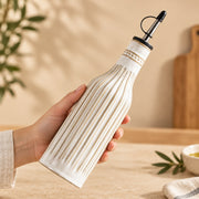 A hand holds a ribbed ceramic oil bottle with a black pour spout against a warm beige kitchen background.