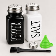 A set of two ceramic shakers, one black labeled 'PEPPER' and one white labeled 'SALT', with a small green funnel, a metal shatter guard, and a black cleaning brush on a light gray background.